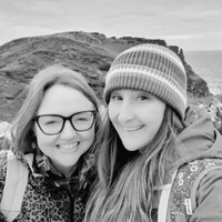 Two women, Ceryn Rowntree (left) and Charlie Edwards (right) stand before a rocky outcrop leading out onto the sea. Both wear raincoats and rucksacks. Ceryn is wearing black rimmed glasses, while Charlie wears a striped woollen hat.
