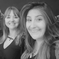 The photograph shows Ceryn Rowntree (left) and Charlie Edwards (right) against a black backdrop, with both smiling at the camera. Charlie is a brunette in her 40s who wears a loose kaftan over a black top and has a bag on the shoulder closest to the camera. Ceryn is a redhead in her 40s wearing a black dress and a large golden necklace. The photo is in black and white.