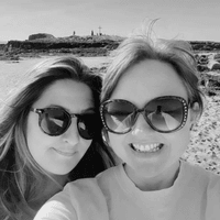 The photograph shows two women, Charlie Edwards (left) and Ceryn Rowntree (right) both standing together by the seaside. Both are white brunettes and wear large sunglasses. The photo is in black and white.