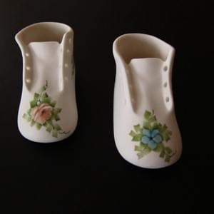 The front toe of two porcelain baby shoes showing the delicately painted flows and foliage. One has a blue forget-me-nota and one has a pink rose.
