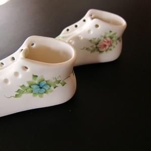 The left hand side of two porcelain baby shoes showing the delicately painted flows and foliage. One has a blue forget-me-nota and one has a pink rose.