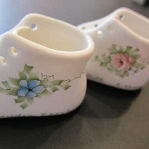The left hand side of two porcelain baby shoes showing the delicately painted flows and foliage. One has a blue forget-me-nota and one has a pink rose.