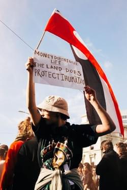 Young man holding a tino rangatiratanga flag and a sign reading "Money doesn't give life. The land does. Protect Ihumātao" Young man holding a tino rangatiratanga flag and a sign reading "Money doesn't give life. The land does. Protect Ihumātao"