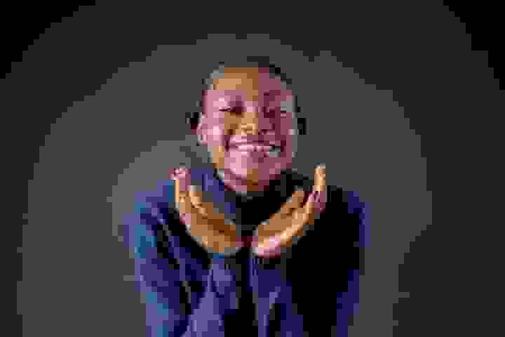A person stands against a dark background, wearing a dark blue turtleneck sweater with their hair styled in two small buns. Their hands are raised near their face, palms upward and close together, fingers gently curved. Focused lighting highlights their face and hands, creating strong contrast with the deep background and giving the portrait a calm, expressive atmosphere. A person stands against a dark background, wearing a dark blue turtleneck sweater with their hair styled in two small buns. Their hands are raised near their face, palms upward and close together, fingers gently curved. Focused lighting highlights their face and hands, creating strong contrast with the deep background and giving the portrait a calm, expressive atmosphere.