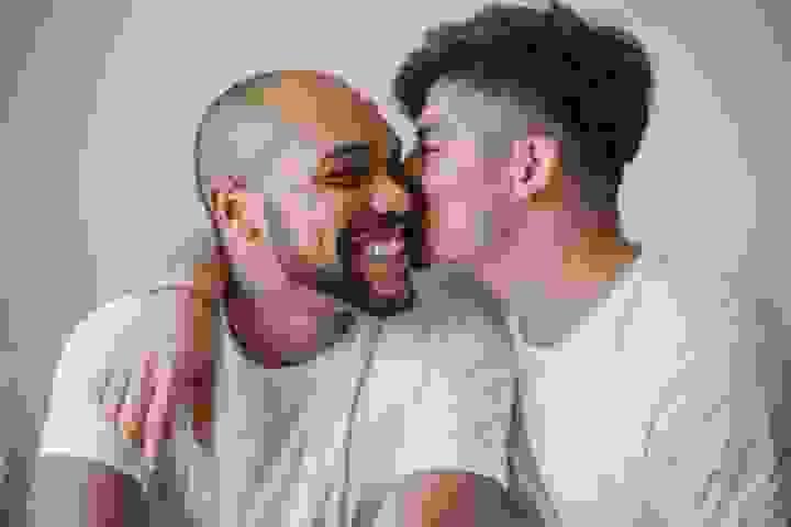 Two people sit close together indoors, both wearing white shirts. One person with short dark hair leans in to kiss the cheek of the other, who has a shaved head, a beard, and a broad smile. Their faces are close, and the moment feels warm, affectionate, and relaxed against a simple neutral background Two people sit close together indoors, both wearing white shirts. One person with short dark hair leans in to kiss the cheek of the other, who has a shaved head, a beard, and a broad smile. Their faces are close, and the moment feels warm, affectionate, and relaxed against a simple neutral background