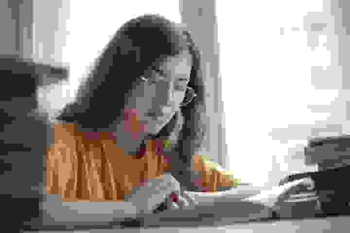 A person with long dark hair and glasses sits at a table reading a book, wearing a yellow shirt. Several books are stacked nearby, and soft natural light comes through a window behind them. The scene feels calm and focused, capturing a quiet moment of concentration and learning A person with long dark hair and glasses sits at a table reading a book, wearing a yellow shirt. Several books are stacked nearby, and soft natural light comes through a window behind them. The scene feels calm and focused, capturing a quiet moment of concentration and learning
