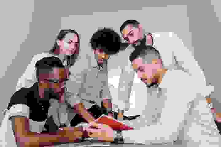 Five people stand closely together indoors, gathered around a red notebook or folder that one of them is holding. They appear to be discussing or reviewing something, leaning in with focused expressions. The setting has neutral walls and bright, even lighting, giving the scene a collaborative, team‑oriented feel. Five people stand closely together indoors, gathered around a red notebook or folder that one of them is holding. They appear to be discussing or reviewing something, leaning in with focused expressions. The setting has neutral walls and bright, even lighting, giving the scene a collaborative, team‑oriented feel.