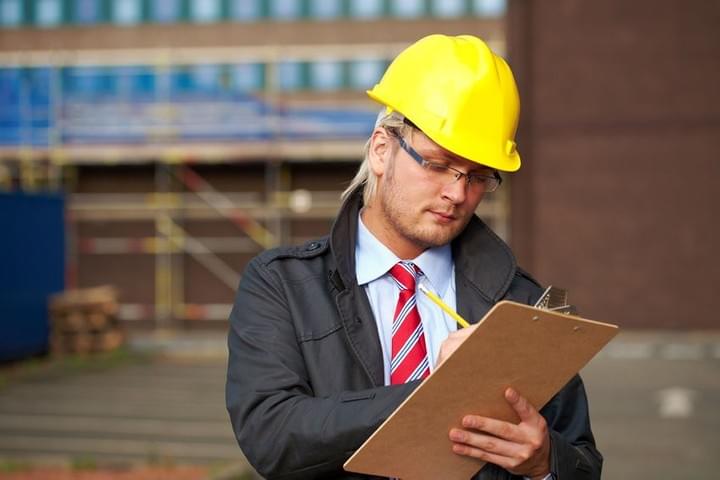 A male access consultant is on a building site, wearing a hardhat and using a clip board to take notes A male access consultant is on a building site, wearing a hardhat and using a clip board to take notes