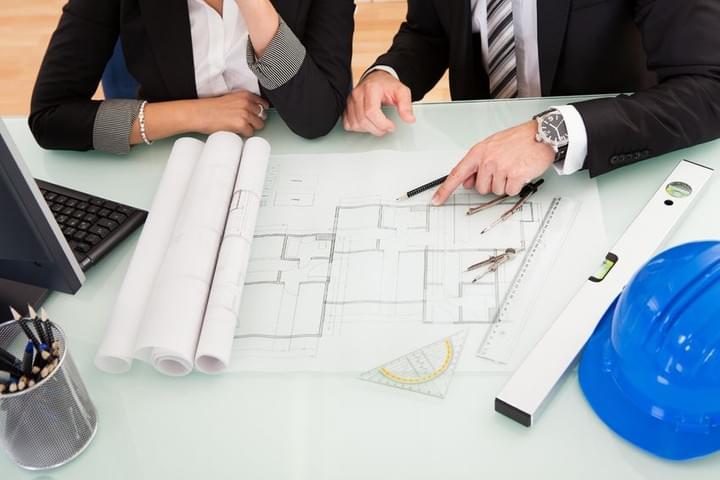 Two people are seated at a desk and are reviewing a set of building plans, the desk also has a laptop, level, and hardhat on it Two people are seated at a desk and are reviewing a set of building plans, the desk also has a laptop, level, and hardhat on it