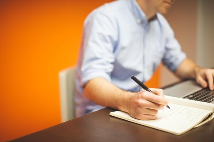 A man is seated at a desk, using a laptop and writing notes into a notebook A man is seated at a desk, using a laptop and writing notes into a notebook