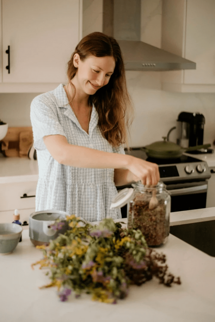 An image of Aleksa, a long haired person wearing a blue and white dress, smiling while preparing a herbal tea in a kitchen. There is a bundle of goldenrod and aster in front of her and a teapot to her right. An image of Aleksa, a long haired person wearing a blue and white dress, smiling while preparing a herbal tea in a kitchen. There is a bundle of goldenrod and aster in front of her and a teapot to her right.