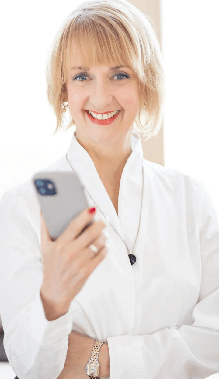 Martina Brandes in weißer Businessbluse hält ein Handy in der rechten Hand und lächelt dich an Martina Brandes in weißer Businessbluse hält ein Handy in der rechten Hand und lächelt dich an