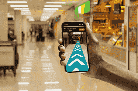 商業施設の通路で、手に持ったスマートフォン画面にARの進行方向ガイドが表示され、目的地までのルートを示している様子。 商業施設の通路で、手に持ったスマートフォン画面にARの進行方向ガイドが表示され、目的地までのルートを示している様子。