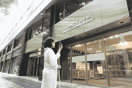 建物の入口前で白杖を持ち、スマートフォンを使って周囲を確認する視覚障害者の様子。 建物の入口前で白杖を持ち、スマートフォンを使って周囲を確認する視覚障害者の様子。