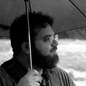 Black and white headshot of Nima Motamed holding an umbrella and looking off camera to the right. Black and white headshot of Nima Motamed holding an umbrella and looking off camera to the right.