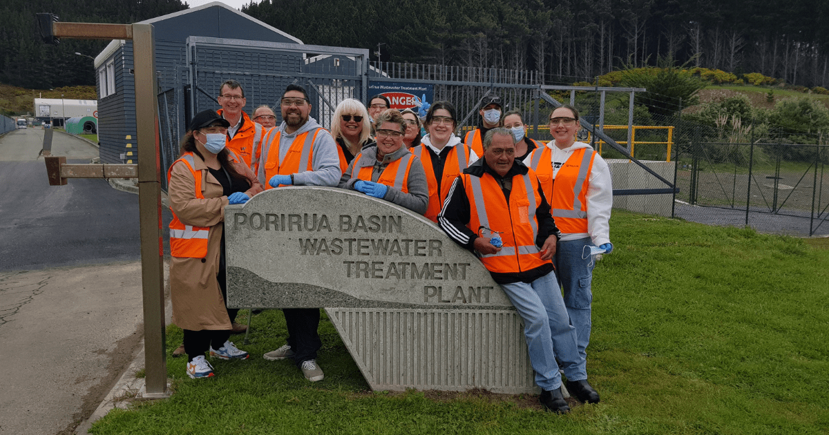 A group of people of different ages, wearing orange high visibility vests, are grouped around a monument that marks the entry to the Porirua basin wastewater treatment plant. A group of people of different ages, wearing orange high visibility vests, are grouped around a monument that marks the entry to the Porirua basin wastewater treatment plant.