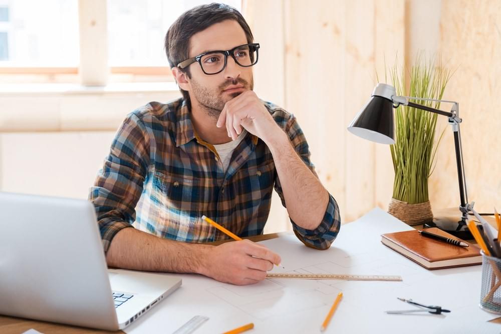 Man in 30s sitting at desk thinking and working on a laptop Man in 30s sitting at desk thinking and working on a laptop
