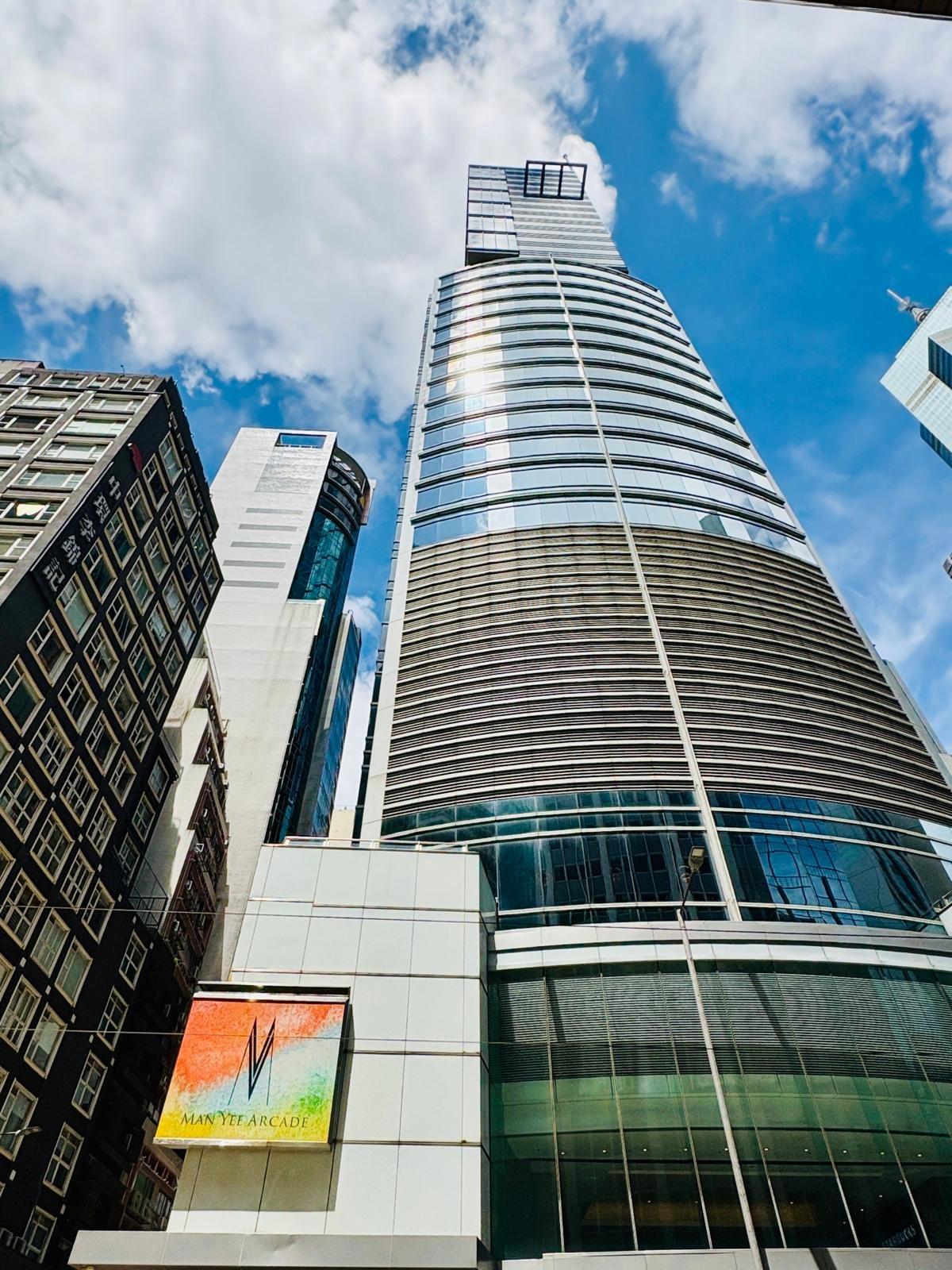 Street-level view looking up at the modern 35-storey glass tower of Man Yee Building, Central Hong Kong, with Man Yee Arcade signage at the base under a bright sky Street-level view looking up at the modern 35-storey glass tower of Man Yee Building, Central Hong Kong, with Man Yee Arcade signage at the base under a bright sky