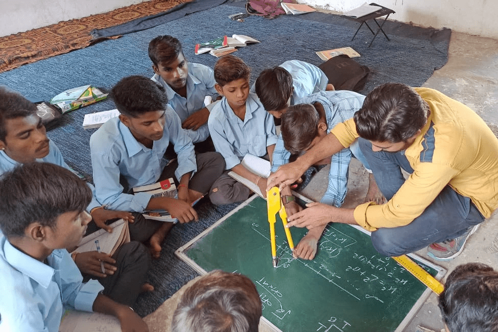 A group of students learning geometry in a maths lesson, sitting on the floor around a diganth facilitator using a large compass. A group of students learning geometry in a maths lesson, sitting on the floor around a diganth facilitator using a large compass.