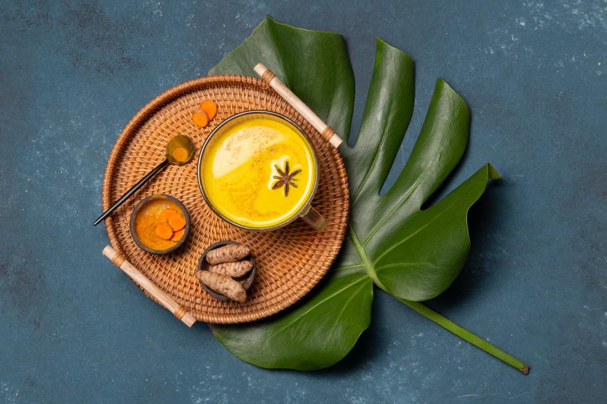 An arrangement featuring raw turmeric, turmeric powder, and a cup of turmeric water garnished with star anise, all presented on a tray placed atop a large green leaf. An arrangement featuring raw turmeric, turmeric powder, and a cup of turmeric water garnished with star anise, all presented on a tray placed atop a large green leaf.