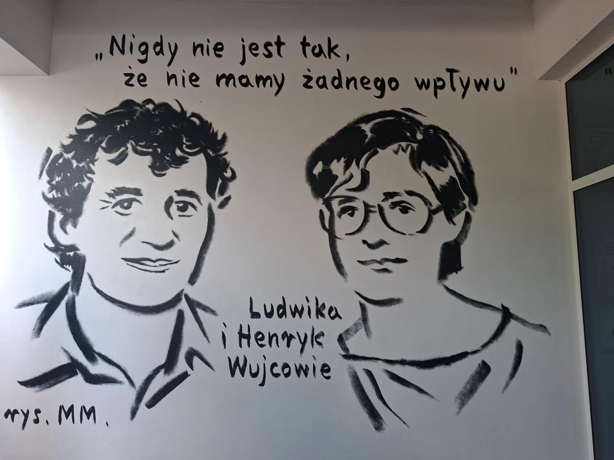 Mężczyzna i kobieta, młodzi.  Głównie twarze i zarysy ramion. On ma kędzierzawe włosy, ona ktrótko obcięte, nosi okulary. Nad nimi napis "Nigdy nie jest tak, że nie mamy żadnego wpływu". Pomiędzy nimi napis "Ludwika i Henryk Wujcowie".  W lewym dolnym rogu podpis rys.  MM. Mężczyzna i kobieta, młodzi.  Głównie twarze i zarysy ramion. On ma kędzierzawe włosy, ona ktrótko obcięte, nosi okulary. Nad nimi napis "Nigdy nie jest tak, że nie mamy żadnego wpływu". Pomiędzy nimi napis "Ludwika i Henryk Wujcowie".  W lewym dolnym rogu podpis rys.  MM.