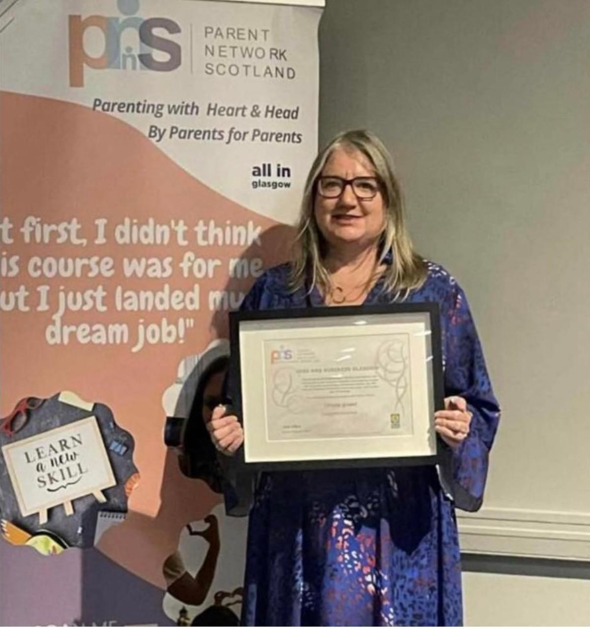 A person stands smiling while holding a framed certificate in front of a Parent Network Scotland banner. A person stands smiling while holding a framed certificate in front of a Parent Network Scotland banner.