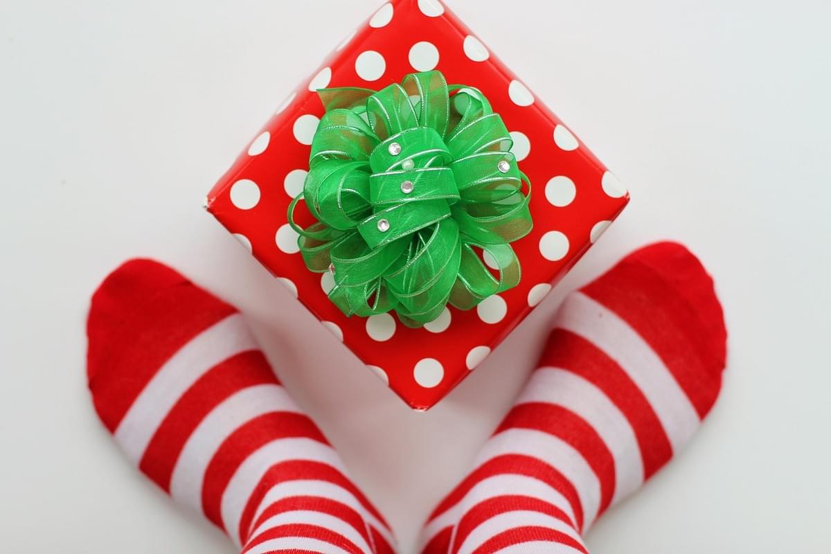 looking down at a box wrapped in red paper with white polka dots and a green bow with little crystals in between two feet wearing striped socks looking down at a box wrapped in red paper with white polka dots and a green bow with little crystals in between two feet wearing striped socks