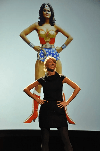 A woman in a black top and fitted skirt stands confidently with her hands on her hips, smiling at the camera. Behind her, a large image of Wonder Woman mirrors her exact pose, wearing her iconic red, blue, and gold costume. The two figures together illustrate the power pose in action A woman in a black top and fitted skirt stands confidently with her hands on her hips, smiling at the camera. Behind her, a large image of Wonder Woman mirrors her exact pose, wearing her iconic red, blue, and gold costume. The two figures together illustrate the power pose in action
