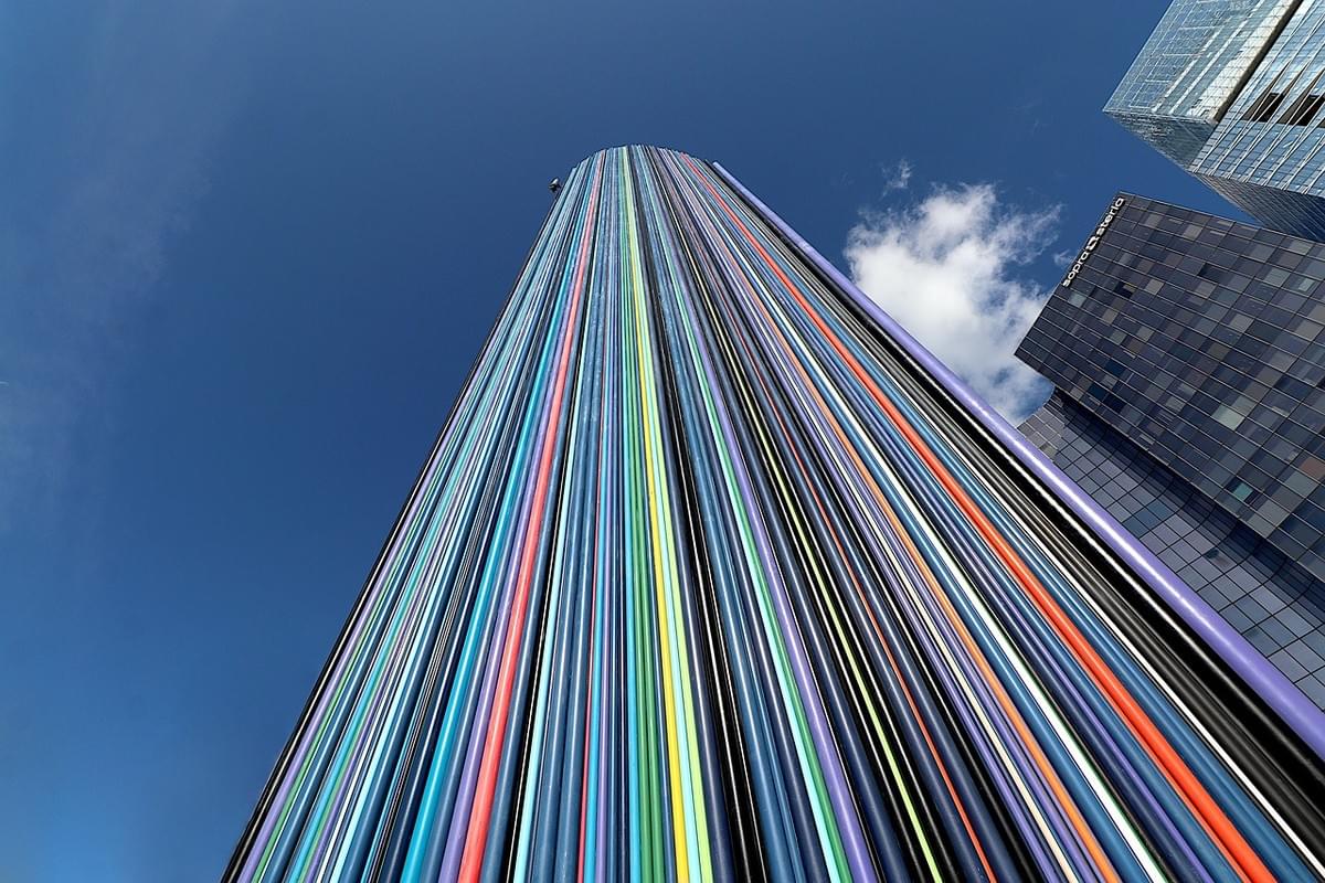 Le Moretti chimney. A chimeny covered in colorful tubes next to some buidlings. The view is looking up and it's against a blue sky with one cloud Le Moretti chimney. A chimeny covered in colorful tubes next to some buidlings. The view is looking up and it's against a blue sky with one cloud