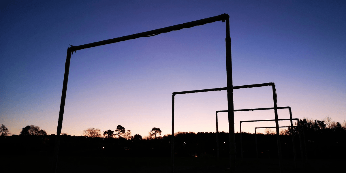 Silhouetted metal frames standing in an open field at twilight, set against a gradient sky shifting from deep blue to soft pink and orange. The geometric structures contrast with the colourful sunset, with distant trees and faint lights visible on the horizon. Silhouetted metal frames standing in an open field at twilight, set against a gradient sky shifting from deep blue to soft pink and orange. The geometric structures contrast with the colourful sunset, with distant trees and faint lights visible on the horizon.