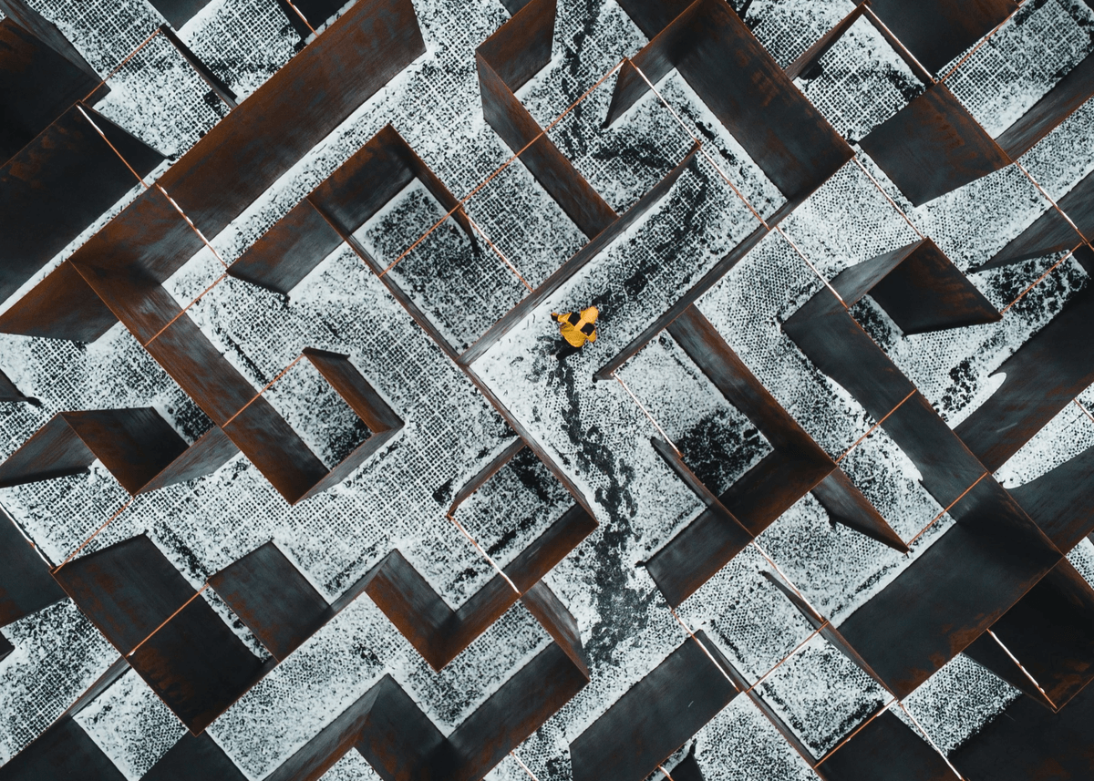 An aerial view of a large outdoor metal maze covered in snow, with a person in a bright yellow jacket walking through the narrow pathways. The rust‑coloured walls, winter setting, and high‑contrast figure create a striking, geometric labyrinth scene. An aerial view of a large outdoor metal maze covered in snow, with a person in a bright yellow jacket walking through the narrow pathways. The rust‑coloured walls, winter setting, and high‑contrast figure create a striking, geometric labyrinth scene.