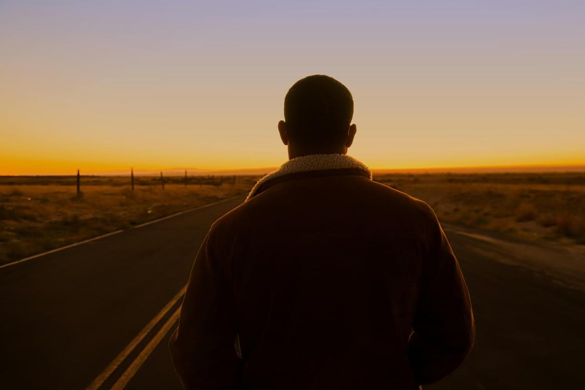 A person in a brown shearling‑collared jacket stands on an empty road at sunset, facing away from the camera. The sky glows with warm orange and purple tones over a sparse desert landscape. A person in a brown shearling‑collared jacket stands on an empty road at sunset, facing away from the camera. The sky glows with warm orange and purple tones over a sparse desert landscape.