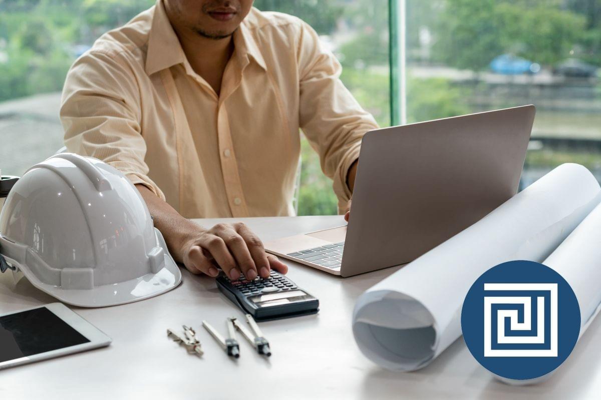 A male architect is looking at a laptop, while using a calculator to check a ramp gradient, there are architectural drawings and a hardhat on the desk A male architect is looking at a laptop, while using a calculator to check a ramp gradient, there are architectural drawings and a hardhat on the desk
