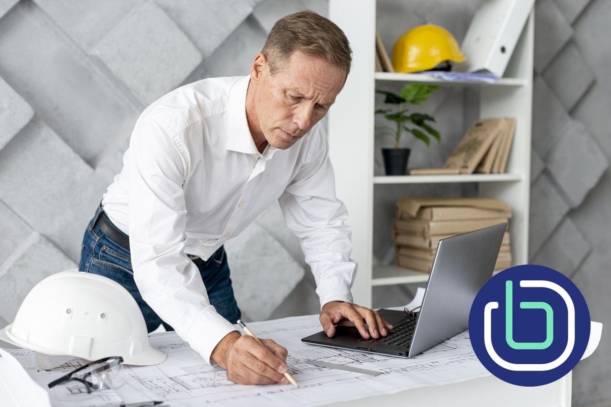 A male architect is looking at a laptop and marking up printed architectural drawings on the desk A male architect is looking at a laptop and marking up printed architectural drawings on the desk