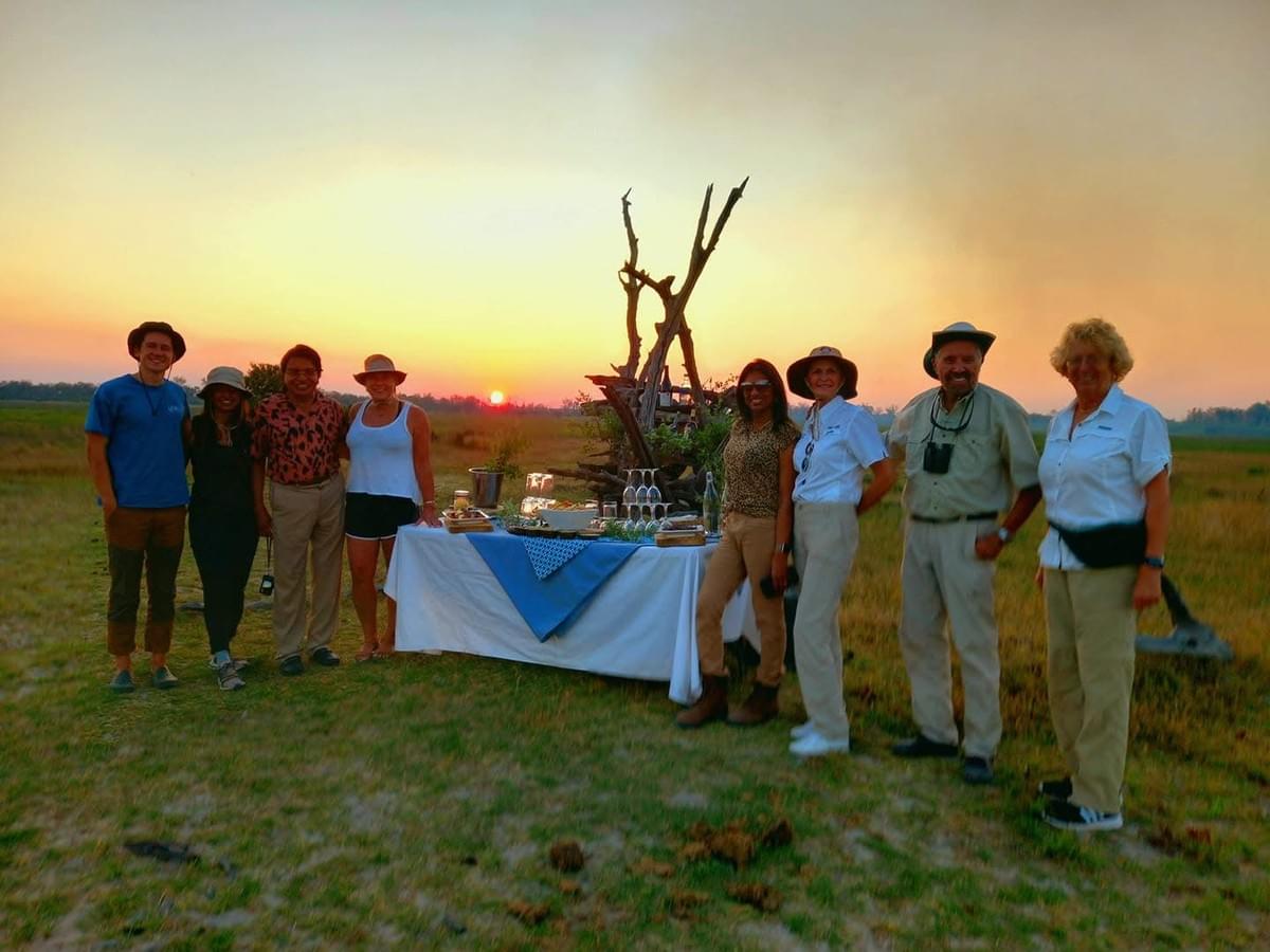Tourists enjoying a sundowner in the middle of the bush, gathered around a table with drinks at sunset. Tourists enjoying a sundowner in the middle of the bush, gathered around a table with drinks at sunset.