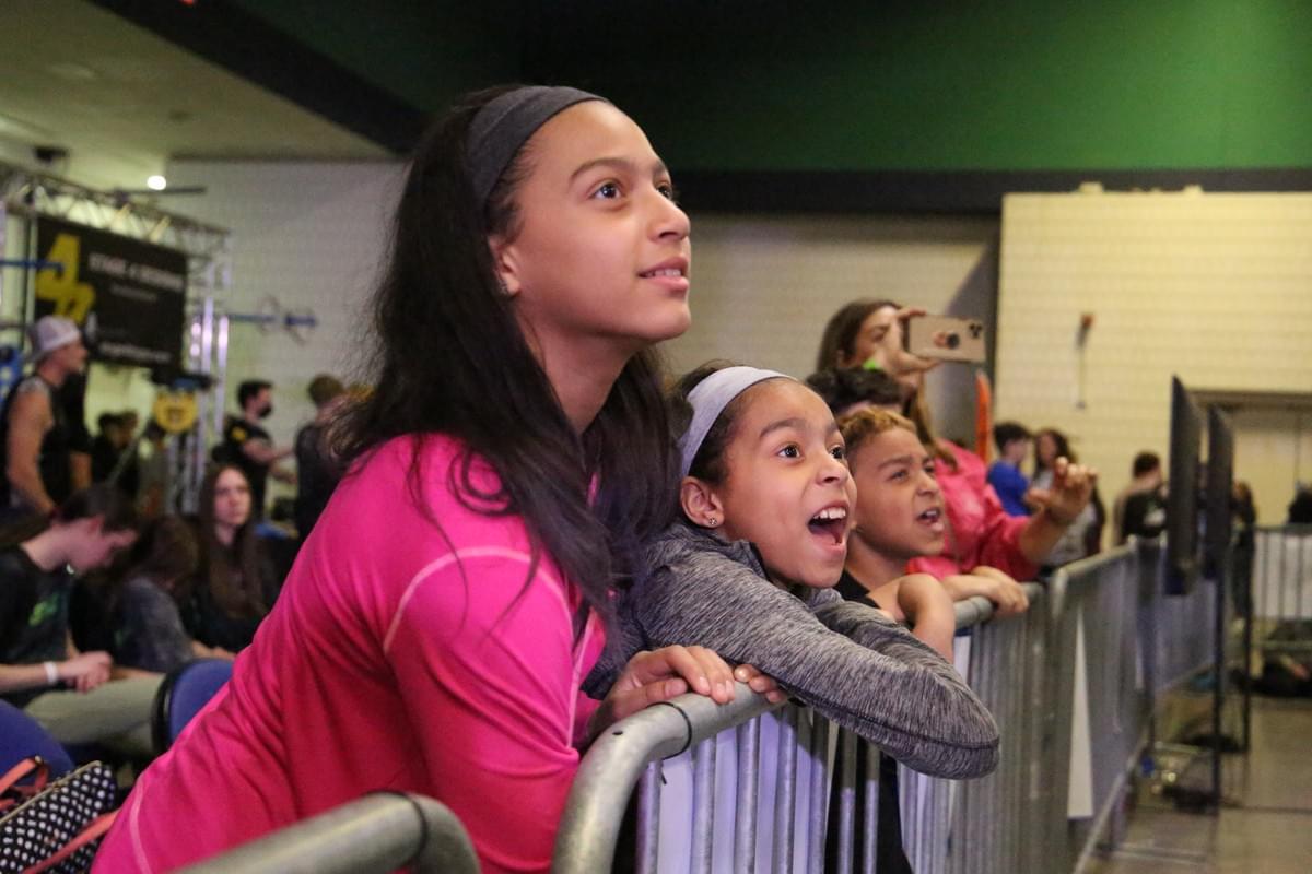 Young StricklyNinja athletes excitedly look on at ninja competitors from behind a metal partition Young StricklyNinja athletes excitedly look on at ninja competitors from behind a metal partition