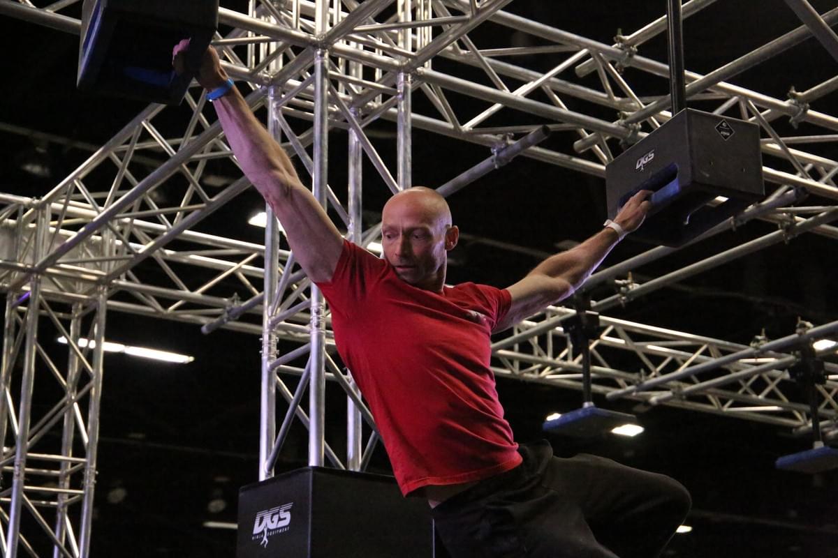 An Adult StricklyNinja is hanging on in mid air to obstacle handles on a ninja course  An Adult StricklyNinja is hanging on in mid air to obstacle handles on a ninja course