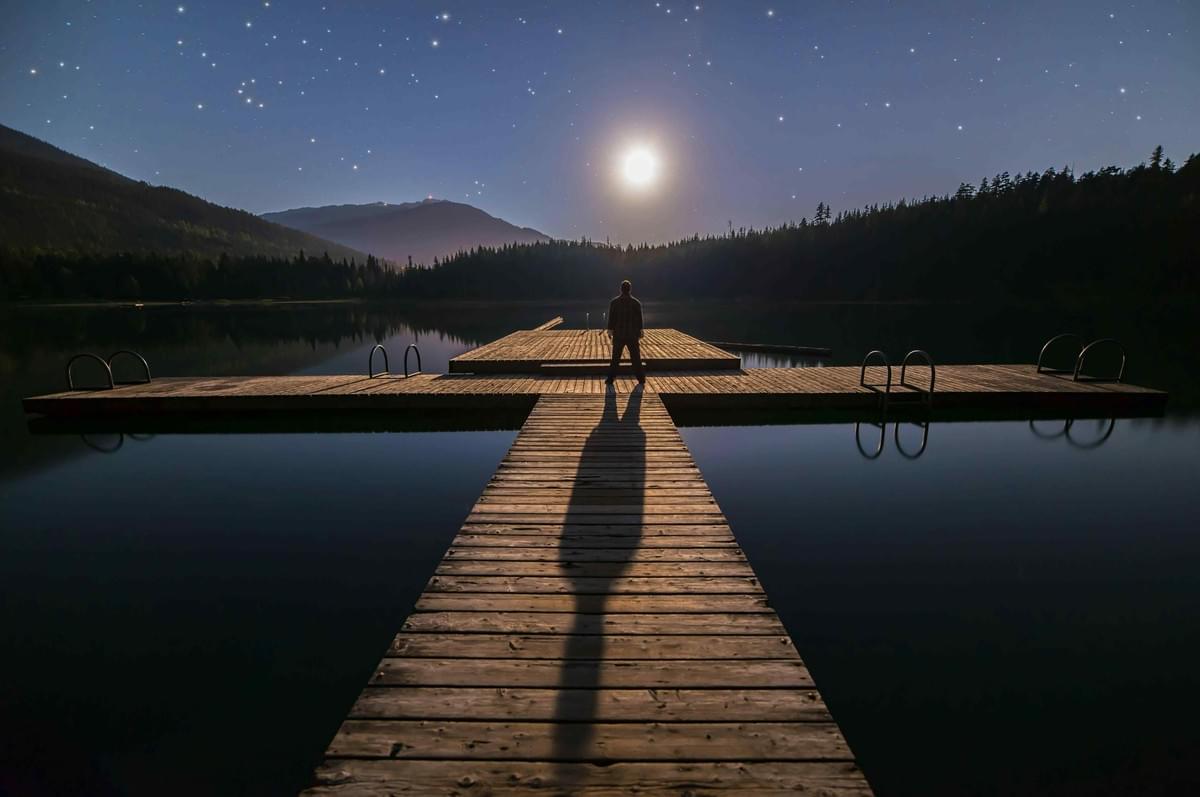 A person standing under the moonglight in the middle of a large finger of docks. The swim ladders and trees appear as silhouettes. A person standing under the moonglight in the middle of a large finger of docks. The swim ladders and trees appear as silhouettes.