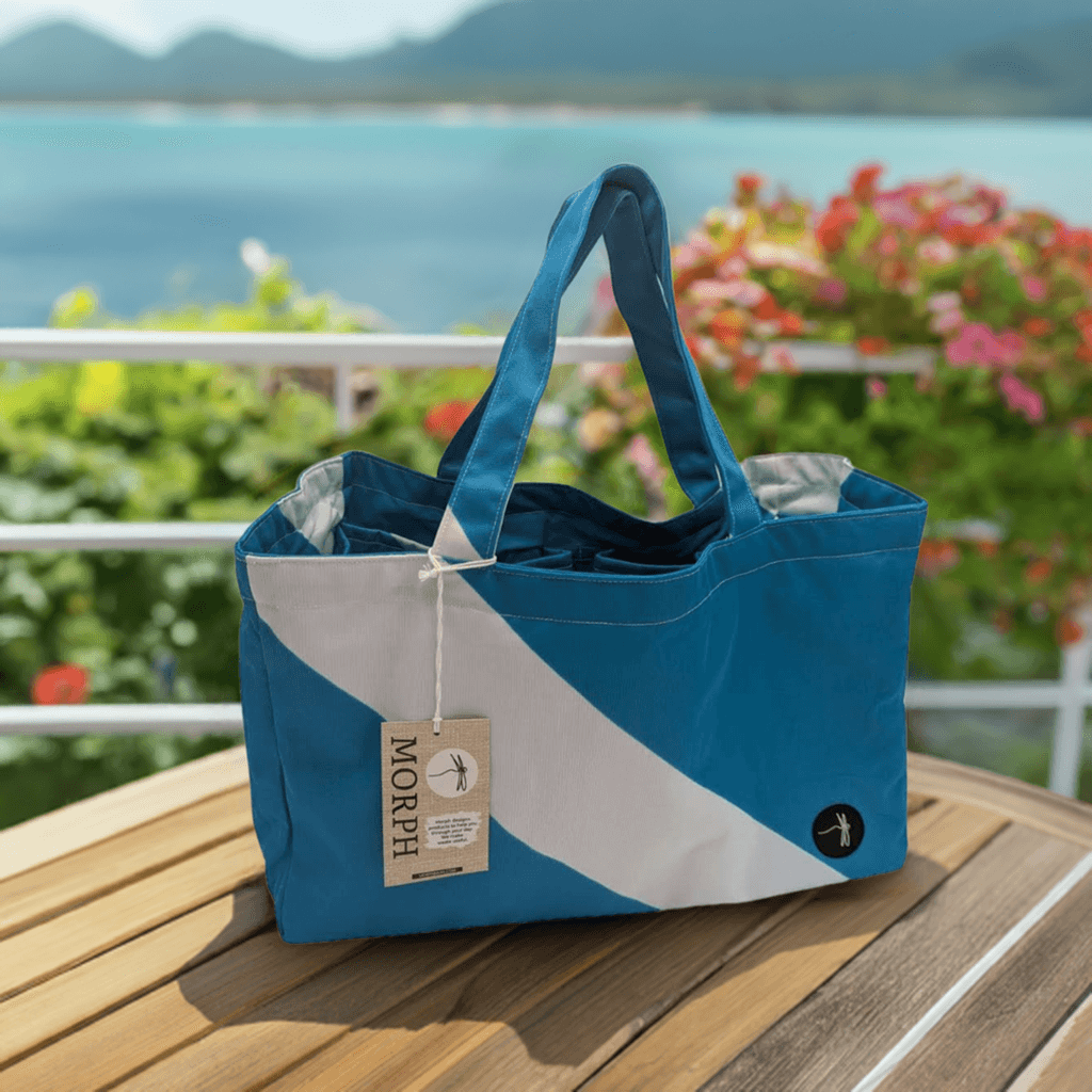 Morph's Market Bag made from upcycled materials, sitting on a round gray table in front of a wooden railing, with the sea visible in the background. Morph's Market Bag made from upcycled materials, sitting on a round gray table in front of a wooden railing, with the sea visible in the background.