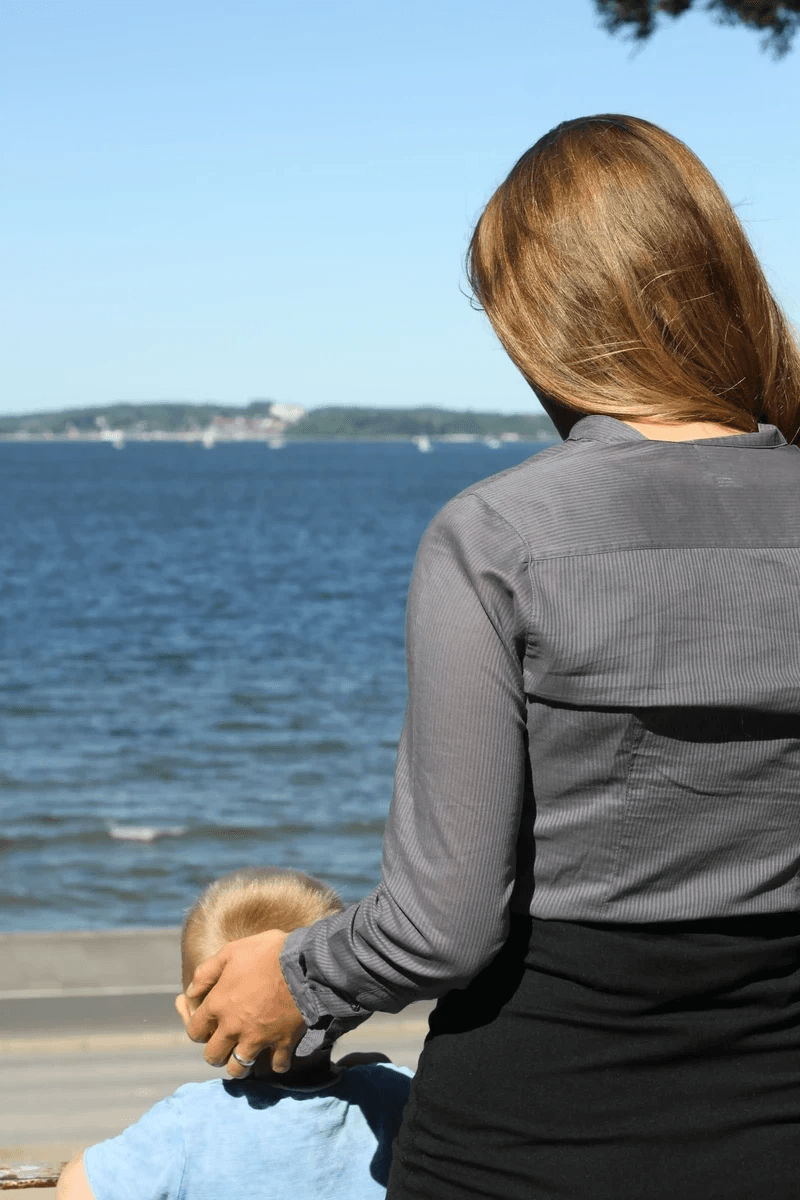 Murena Overhoff steht von hinten zu sehen am Ufer des Wassers, blickt hinaus aufs Wasser und hält schützend ein Kind an ihrer Seite. Murena Overhoff steht von hinten zu sehen am Ufer des Wassers, blickt hinaus aufs Wasser und hält schützend ein Kind an ihrer Seite.