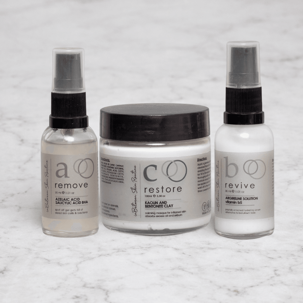 Three In-Between Products add-on range items arranged in a loose triangle on a smooth cool white marble surface with subtle grey veining. Left: small clear glass 30ml mist-spray bottle, black ribbed nozzle with clear overcap, greige matte label, large lowercase serif "a" in dusty mauve, mauve double-circle "OO" logo, "remove" in charcoal serif, "AZELAIC ACID / SALICYLIC ACID BHA" in small caps — near-clear liquid inside. Centre-back: wide squat round glass jar with flat black matte screw lid, greige wrap label, large charcoal "c", neutral charcoal "OO" logo, "restore" in charcoal serif, "KAOLIN AND BENTONITE CLAY" in small caps — white clay contents fully visible through glass sides. Right: identical 30ml spray bottle, greige label, large charcoal "b", "revive" in charcoal, "ARGIRELINE SOLUTION vitamin b5" — white opaque milky contents. Soft cool overhead studio lighting, razor-sharp labels, subtle drop shadows. Ultra-photorealistic, no CGI feel, editorial skincare standard. Three In-Between Products add-on range items arranged in a loose triangle on a smooth cool white marble surface with subtle grey veining. Left: small clear glass 30ml mist-spray bottle, black ribbed nozzle with clear overcap, greige matte label, large lowercase serif "a" in dusty mauve, mauve double-circle "OO" logo, "remove" in charcoal serif, "AZELAIC ACID / SALICYLIC ACID BHA" in small caps — near-clear liquid inside. Centre-back: wide squat round glass jar with flat black matte screw lid, greige wrap label, large charcoal "c", neutral charcoal "OO" logo, "restore" in charcoal serif, "KAOLIN AND BENTONITE CLAY" in small caps — white clay contents fully visible through glass sides. Right: identical 30ml spray bottle, greige label, large charcoal "b", "revive" in charcoal, "ARGIRELINE SOLUTION vitamin b5" — white opaque milky contents. Soft cool overhead studio lighting, razor-sharp labels, subtle drop shadows. Ultra-photorealistic, no CGI feel, editorial skincare standard.