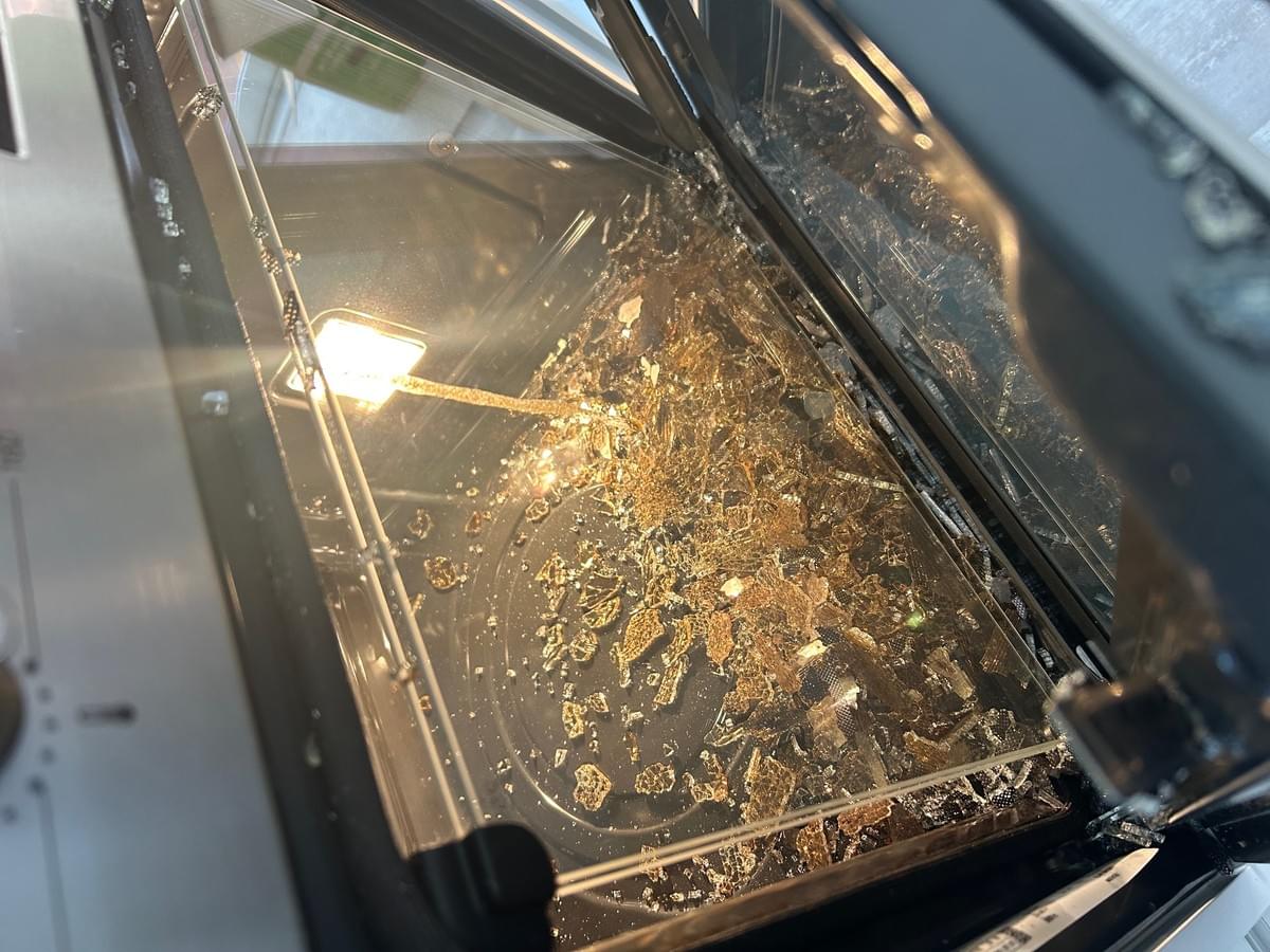 Interior of a oven with shattered glass scattered across the bottom. The broken glass has exploded into small shards. The oven light is on, illuminating the damage. A control dial is partially visible on the left side of the frame. Interior of a oven with shattered glass scattered across the bottom. The broken glass has exploded into small shards. The oven light is on, illuminating the damage. A control dial is partially visible on the left side of the frame.