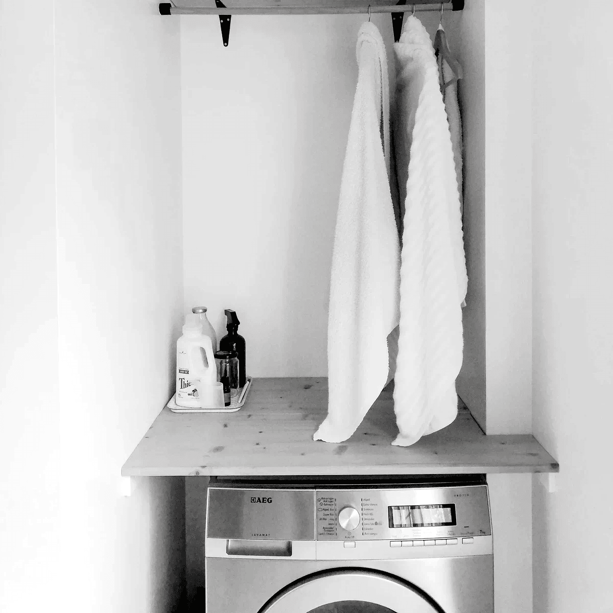 A black and white laundry with a machine machine on the bottom and a wooden shelve holding soaps and detergents with towels and clothes hanging from the top shelf A black and white laundry with a machine machine on the bottom and a wooden shelve holding soaps and detergents with towels and clothes hanging from the top shelf