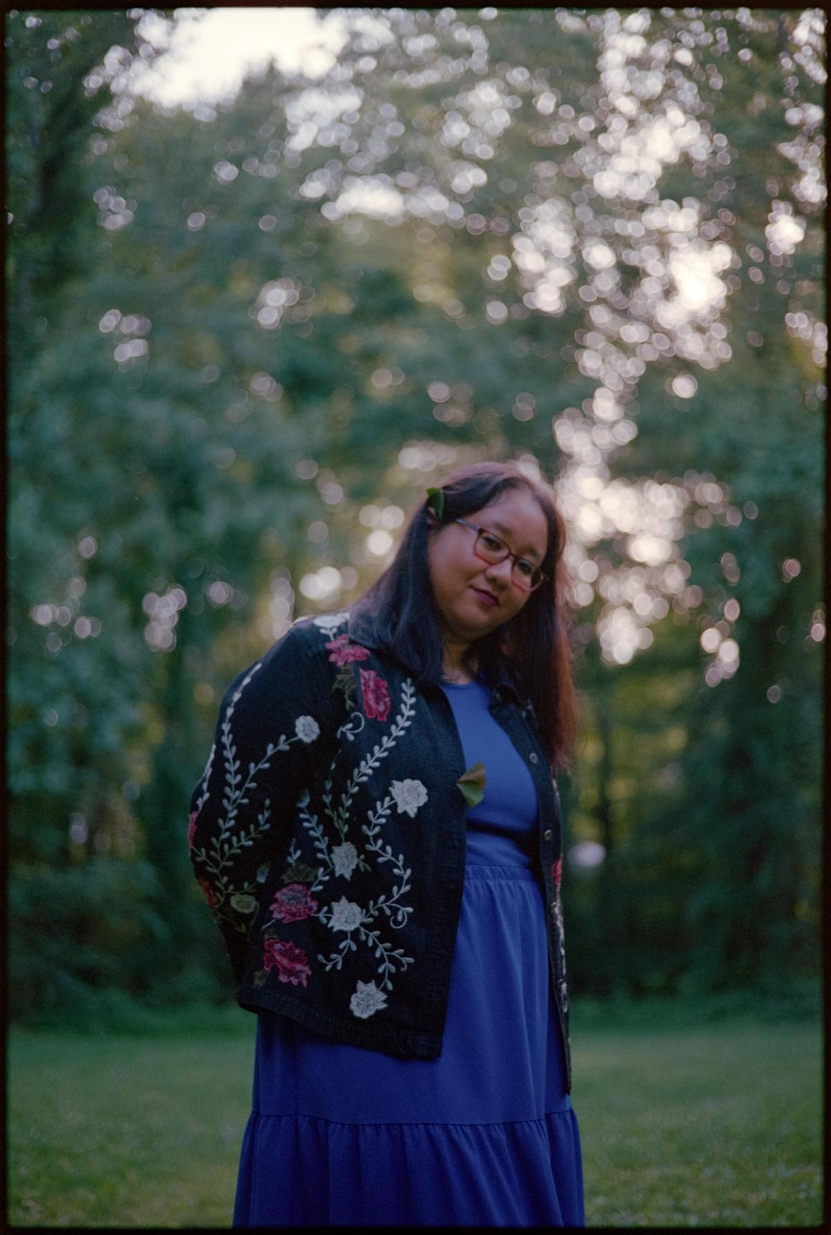 The poet, in a blue dress, standing amid the trees. Photo by Grey Kenna. The poet, in a blue dress, standing amid the trees. Photo by Grey Kenna.