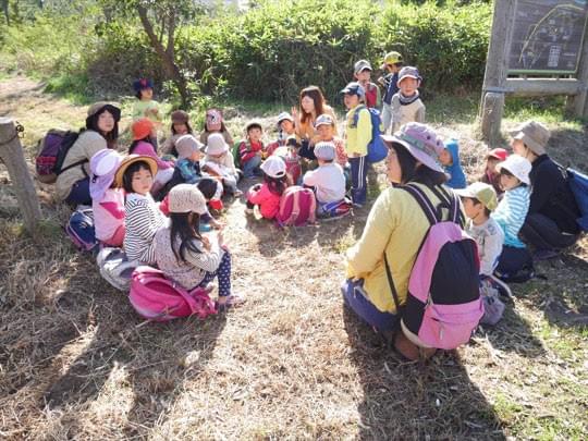 保育者と一緒に森で座って話を聞く子どもたちの写真 保育者と一緒に森で座って話を聞く子どもたちの写真
