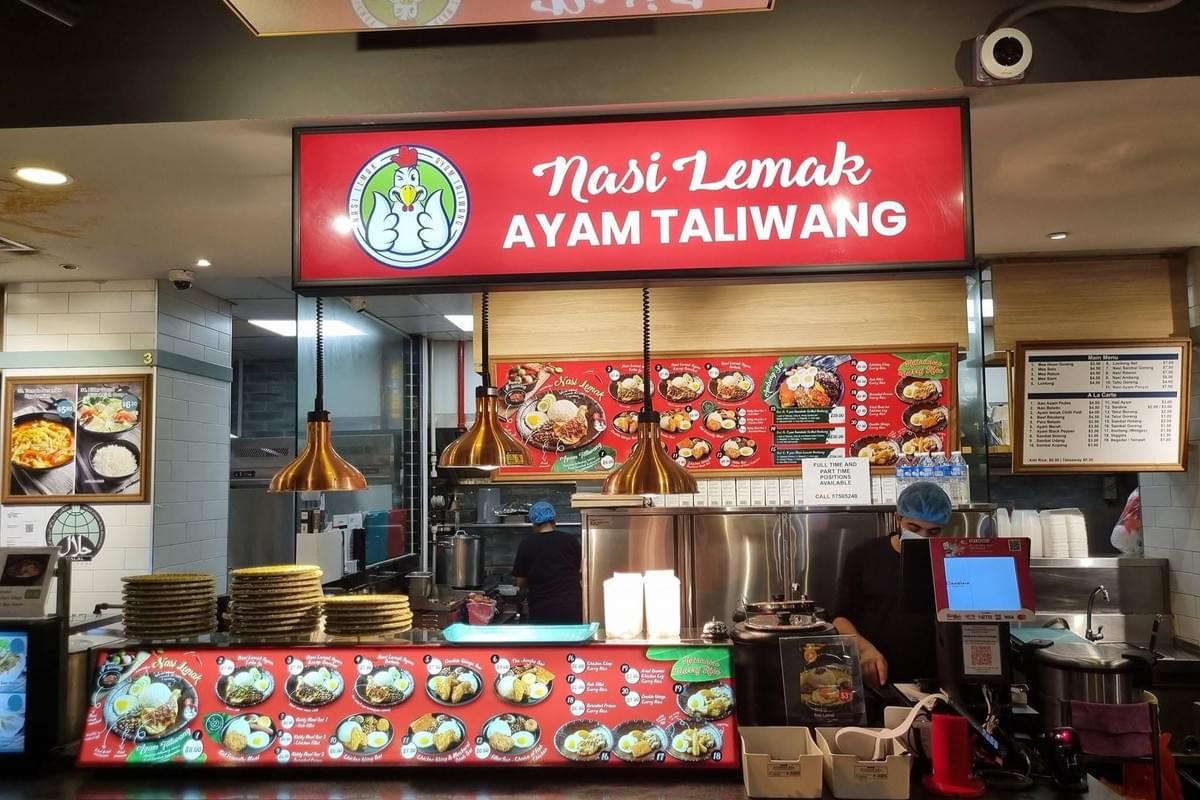A food stall counter displaying a prominent red illuminated sign that reads "Nasi Lemak AYAM TALIWANG" alongside a cartoon chicken mascot. Illuminated menu boards feature photos of various rice dishes, with warm pendant lights hanging over the counter where a worker wearing a face mask stands. A food stall counter displaying a prominent red illuminated sign that reads "Nasi Lemak AYAM TALIWANG" alongside a cartoon chicken mascot. Illuminated menu boards feature photos of various rice dishes, with warm pendant lights hanging over the counter where a worker wearing a face mask stands.