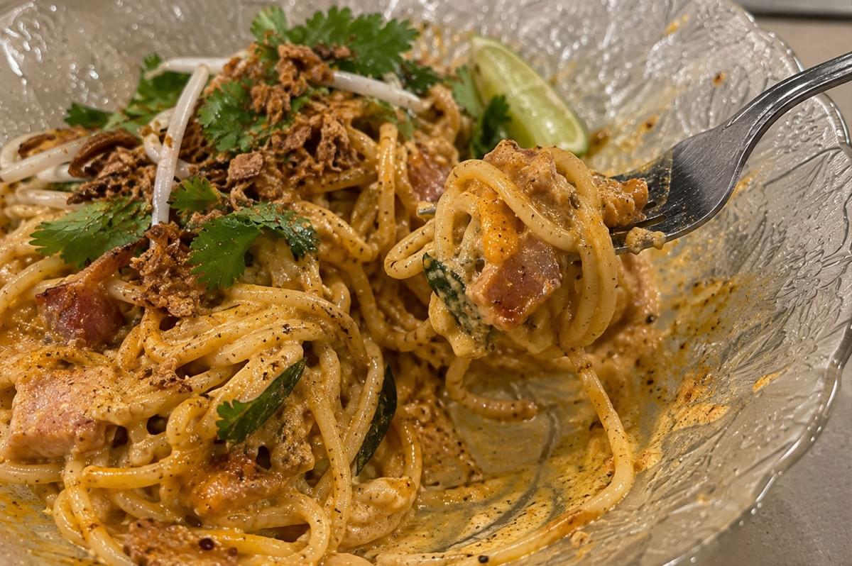 A close-up shot captures a fork twirling a bite of creamy laksa carbonara pasta containing pancetta bits out of a clear textured glass bowl, topped with fried shallots, cilantro, and bean sprouts. A close-up shot captures a fork twirling a bite of creamy laksa carbonara pasta containing pancetta bits out of a clear textured glass bowl, topped with fried shallots, cilantro, and bean sprouts.