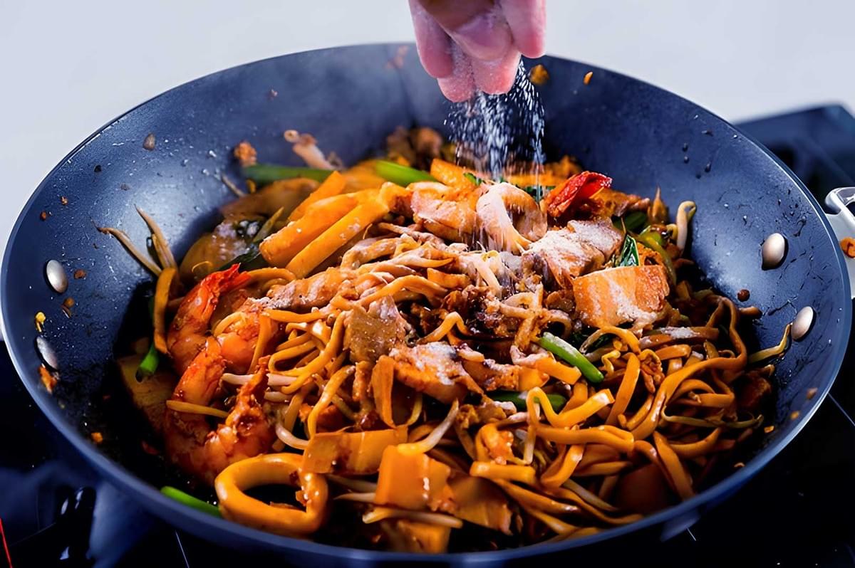 A hand sprinkles salt over sizzling noodles with shrimp and vegetables in a pan, creating a savory, appetizing dish filled with vibrant colors. Hokkien wok A hand sprinkles salt over sizzling noodles with shrimp and vegetables in a pan, creating a savory, appetizing dish filled with vibrant colors. Hokkien wok
