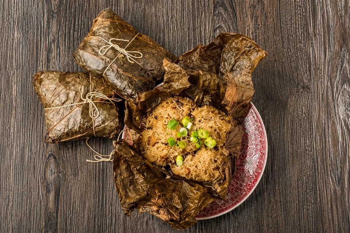 A top-down view of seasoned sticky rice topped with green onions, served in an unwrapped lotus leaf on a patterned plate, alongside two tied leaf parcels on a dark wooden surface. A top-down view of seasoned sticky rice topped with green onions, served in an unwrapped lotus leaf on a patterned plate, alongside two tied leaf parcels on a dark wooden surface.