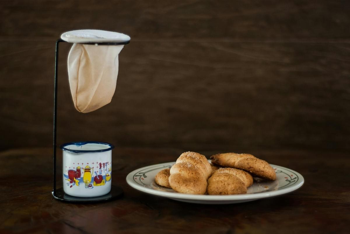 A mini cloth coffee filter strainer stands over a small patterned enamel mug on a dark wooden surface. Next to it, a ceramic plate holds a stack of fresh, baked pastries sprinkled with sugar. A mini cloth coffee filter strainer stands over a small patterned enamel mug on a dark wooden surface. Next to it, a ceramic plate holds a stack of fresh, baked pastries sprinkled with sugar.